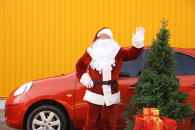 Authentic Santa Claus near car with fir tree and presents against yellow background