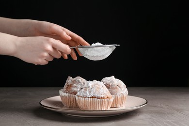 Photo of Woman with sieve sprinkling powdered sugar onto muffins at grey textured table, closeup