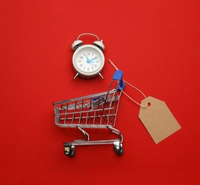 Photo of Shopping cart with tag and alarm clock on red background, flat lay. Sale concept