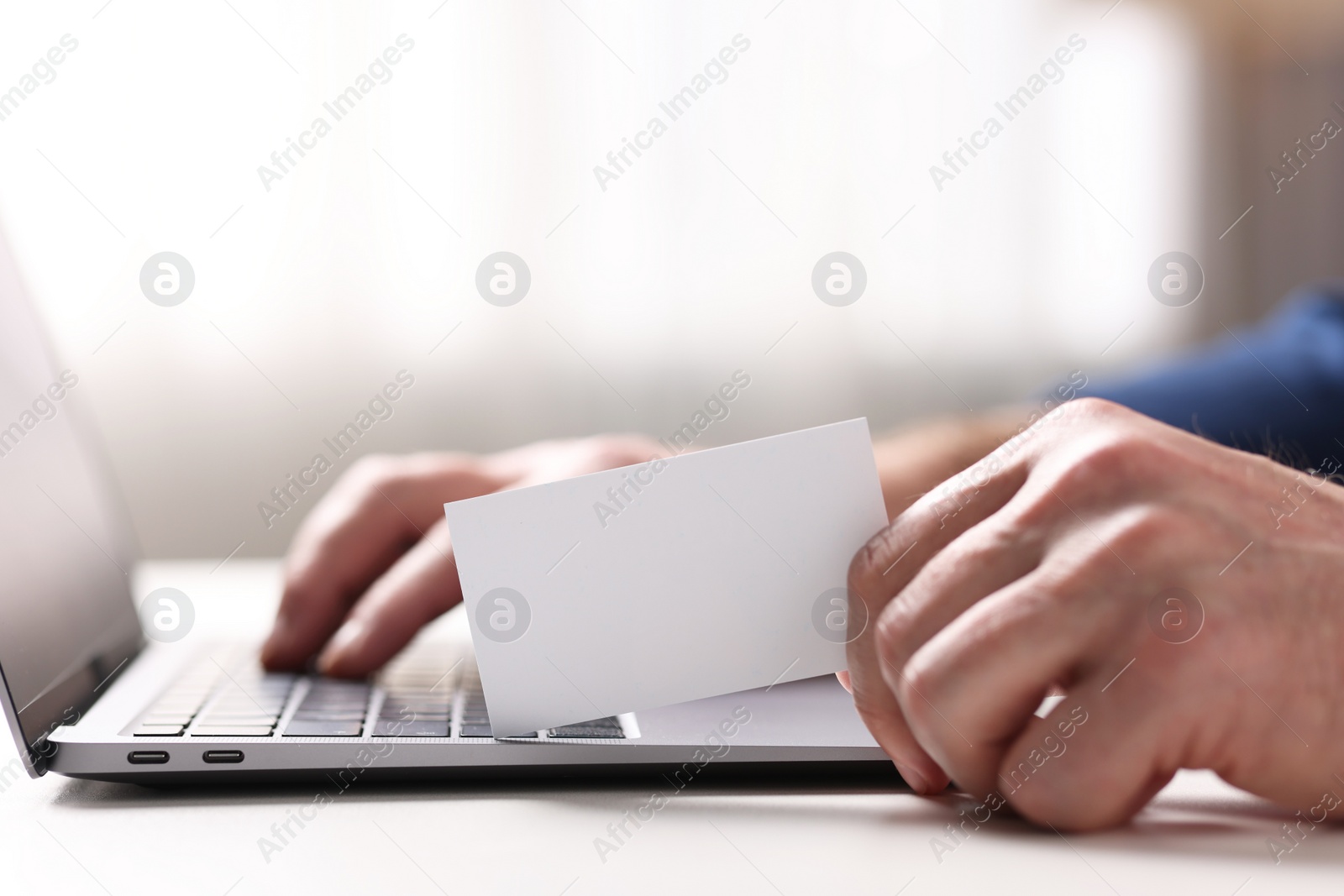 Photo of Man with laptop holding blank business card at white table indoors, closeup