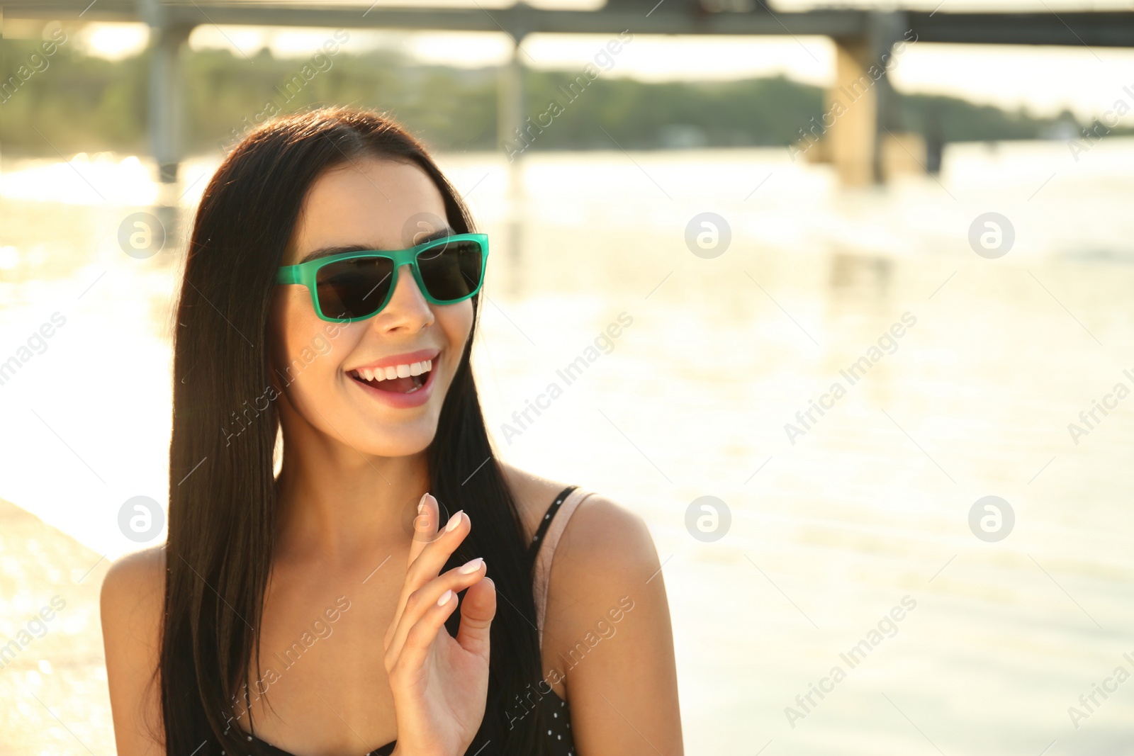 Photo of Beautiful young woman wearing stylish sunglasses near river. Space for text