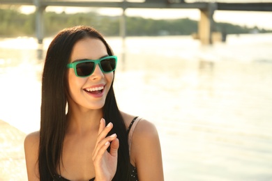 Photo of Beautiful young woman wearing stylish sunglasses near river. Space for text