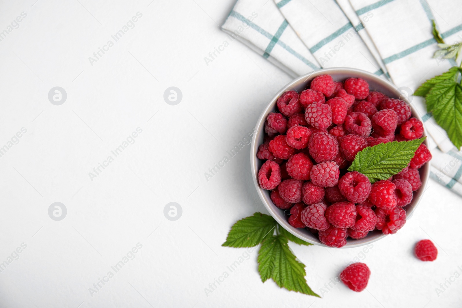 Photo of Bowl with fresh ripe raspberries and green leaves on white table, flat lay. Space for text