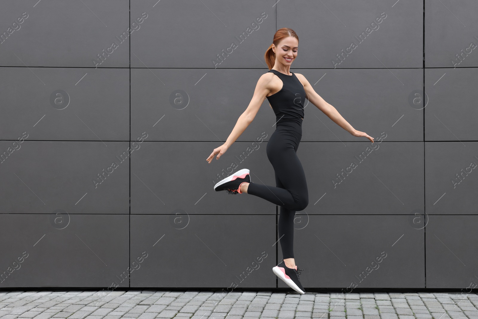 Photo of Beautiful woman in gym clothes jumping near dark grey wall on street