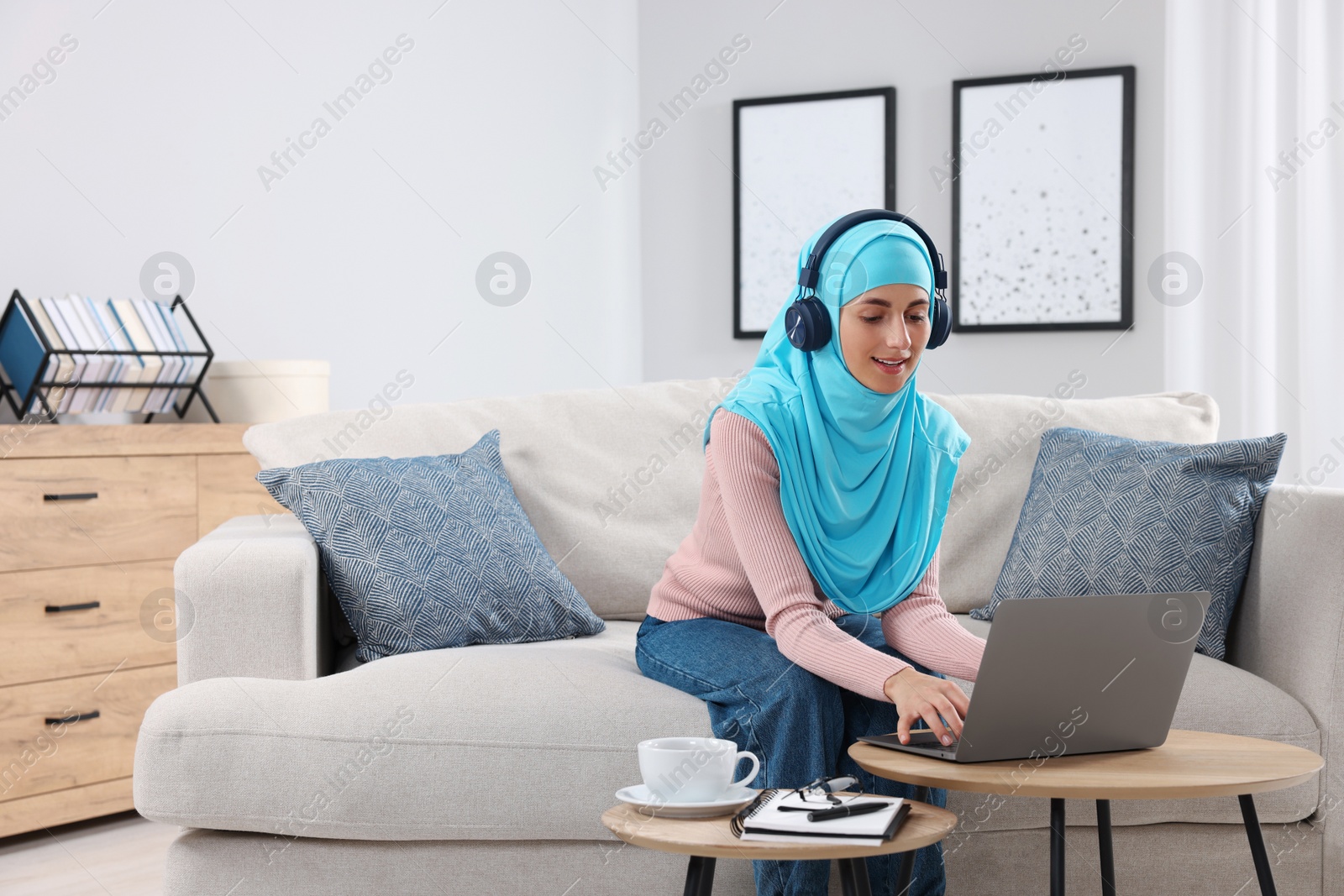 Photo of Muslim woman in headphones using laptop at wooden table in room. Space for text