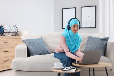 Muslim woman in headphones using laptop at wooden table in room. Space for text