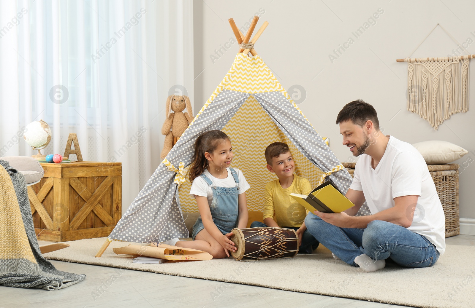 Photo of Father reading book to children near toy wigwam at home