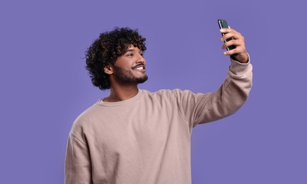 Handsome smiling man taking selfie with smartphone on violet background, space for text