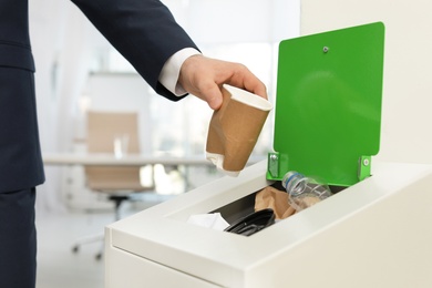 Man putting used paper cup into trash bin in office, closeup. Waste recycling