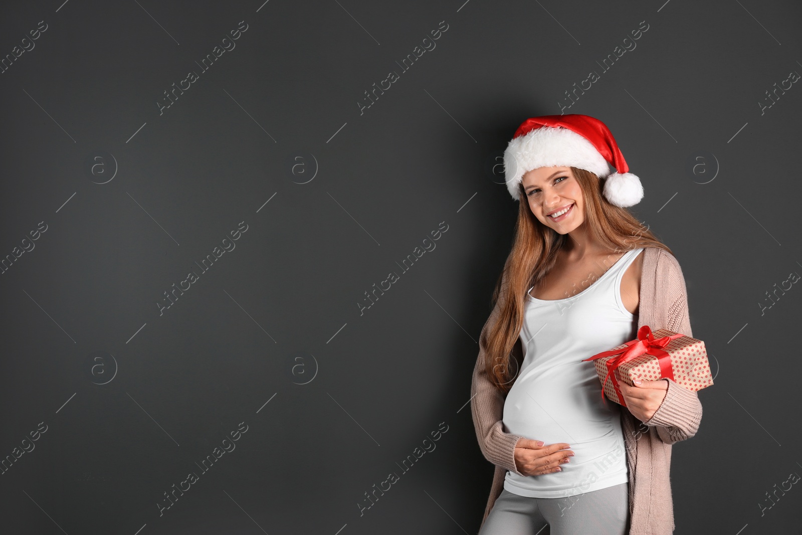 Photo of Young pregnant woman in Santa hat with Christmas gift on grey background, space for text