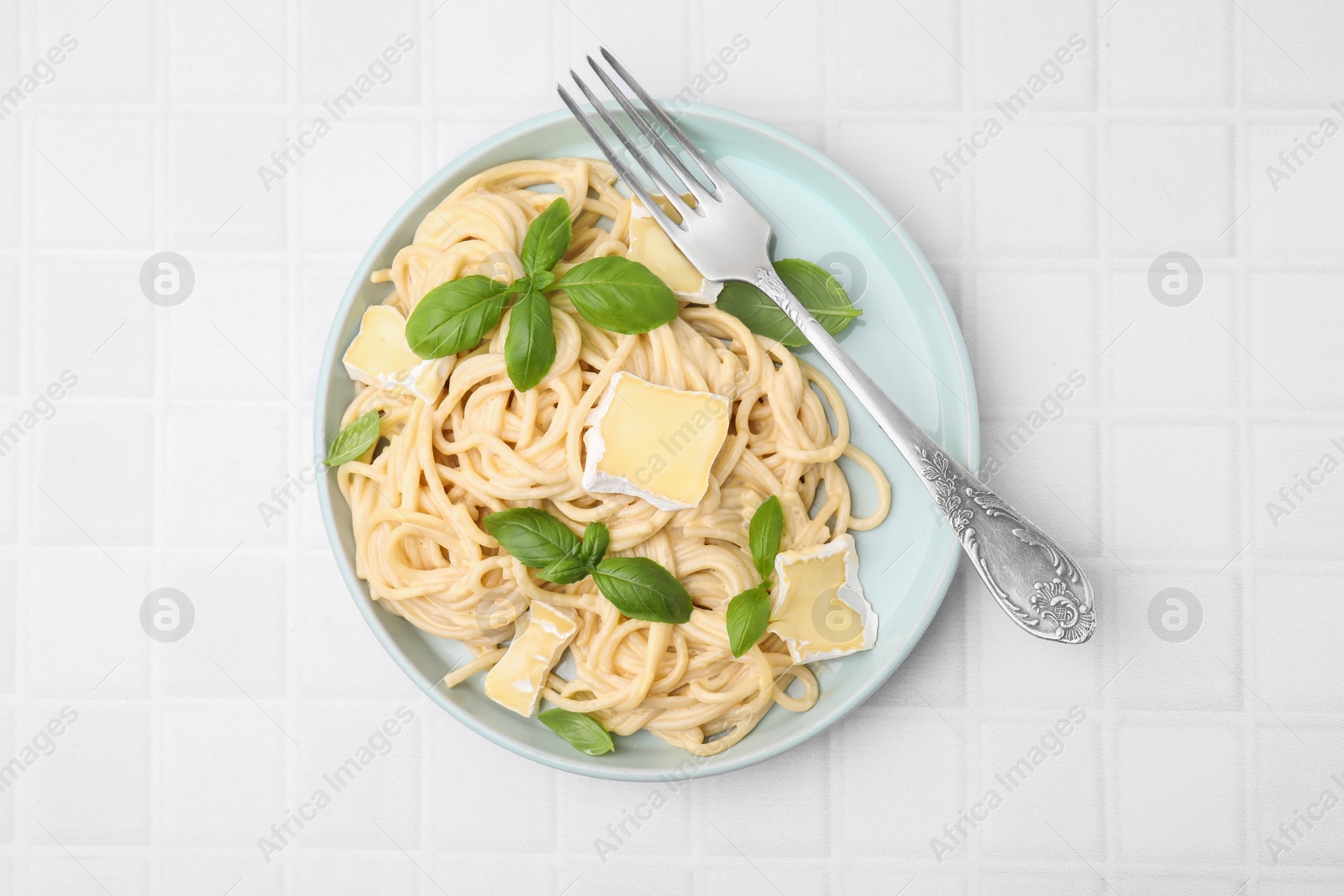 Photo of Delicious pasta with brie cheese, basil and fork on white tiled table, top view