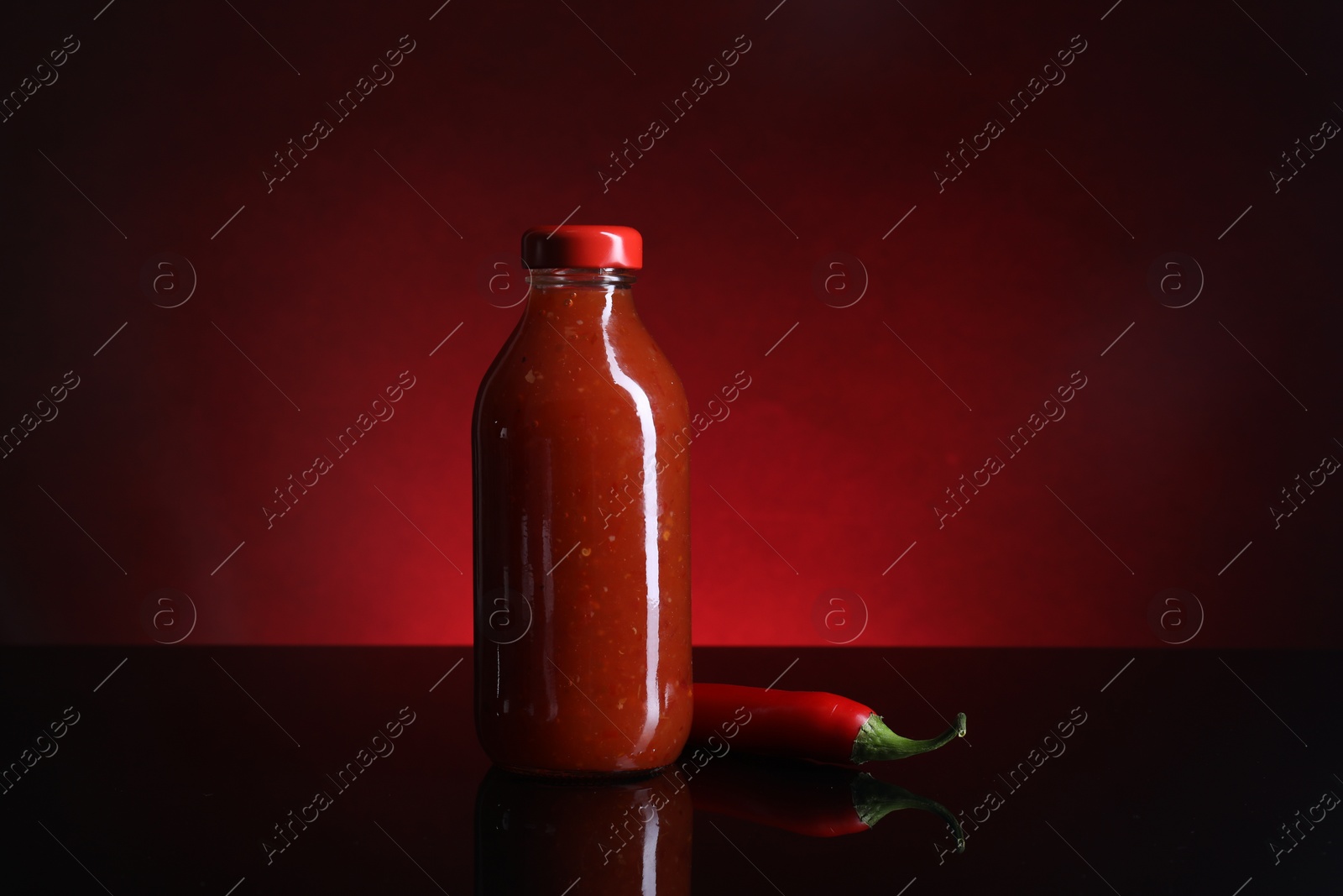 Photo of Spicy chili sauce in bottle and pepper against dark background