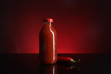 Photo of Spicy chili sauce in bottle and pepper against dark background