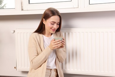 Woman holding cup with hot drink near heating radiator indoors