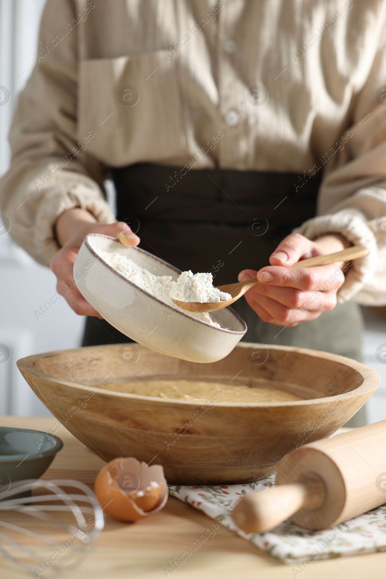 Photo of Making dough. Woman adding flour into bowl at wooden table in kitchen, closeup