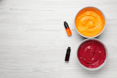 Photo of Bowls of different cream with food coloring and bottles of bright liquid on white wooden table, flat lay. Space for text