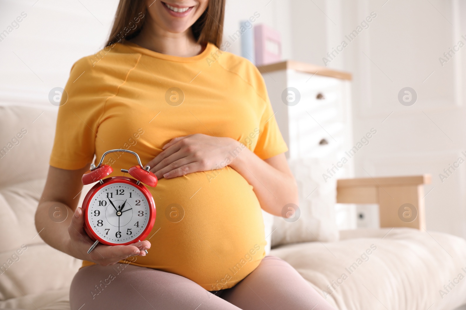 Photo of Young pregnant woman holding alarm clock near her belly at home, closeup. Time to give birth