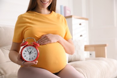 Young pregnant woman holding alarm clock near her belly at home, closeup. Time to give birth