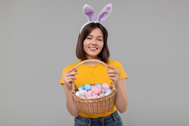 Easter celebration. Happy woman with bunny ears and wicker basket full of painted eggs on grey background