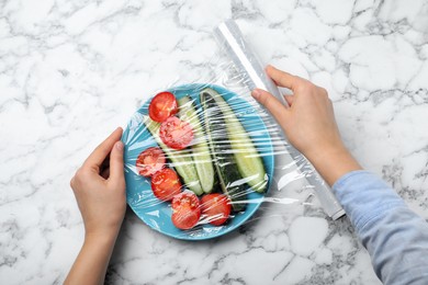 Photo of Woman putting plastic food wrap over plate of fresh vegetables at white marble table, top view