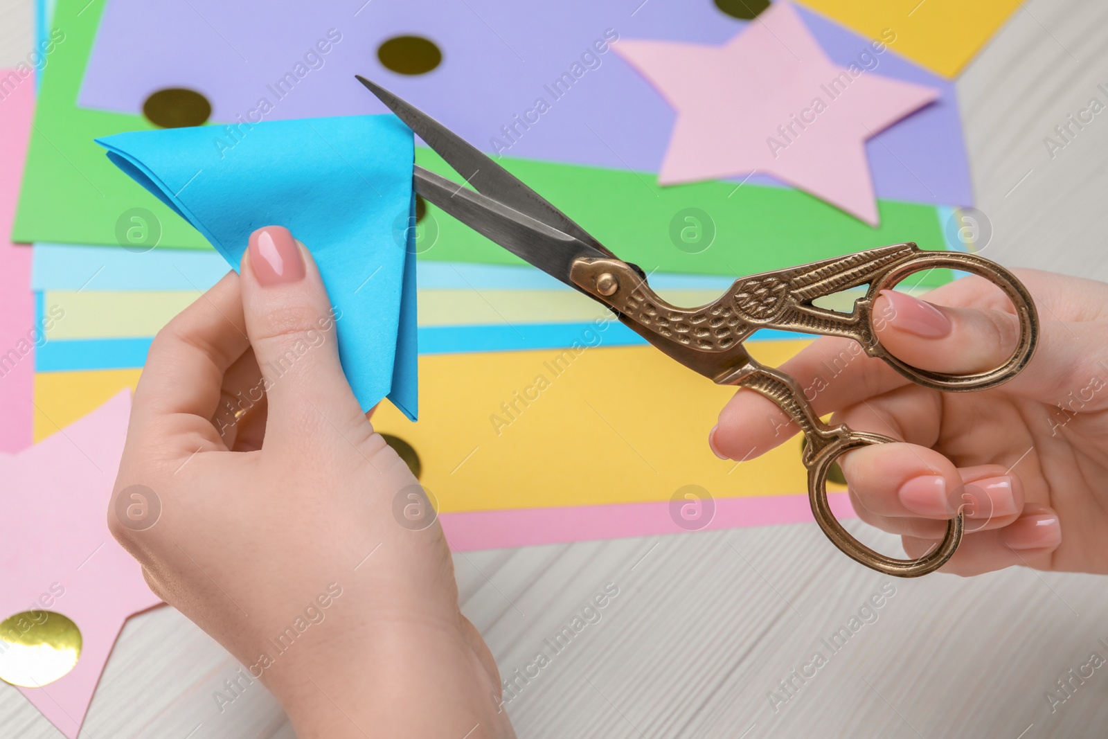 Photo of Woman cutting light blue paper with scissors at white wooden table, closeup