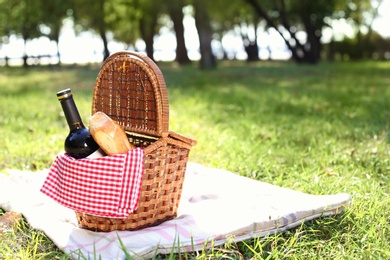 Wicker picnic basket with bottle of wine and bread on blanket in park. Space for text