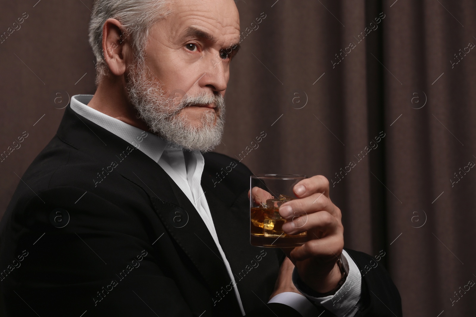 Photo of Senior man in suit holding glass of whiskey with ice cubes on brown background. Space for text