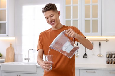 Photo of Happy man pouring water from filter jug into glass in kitchen