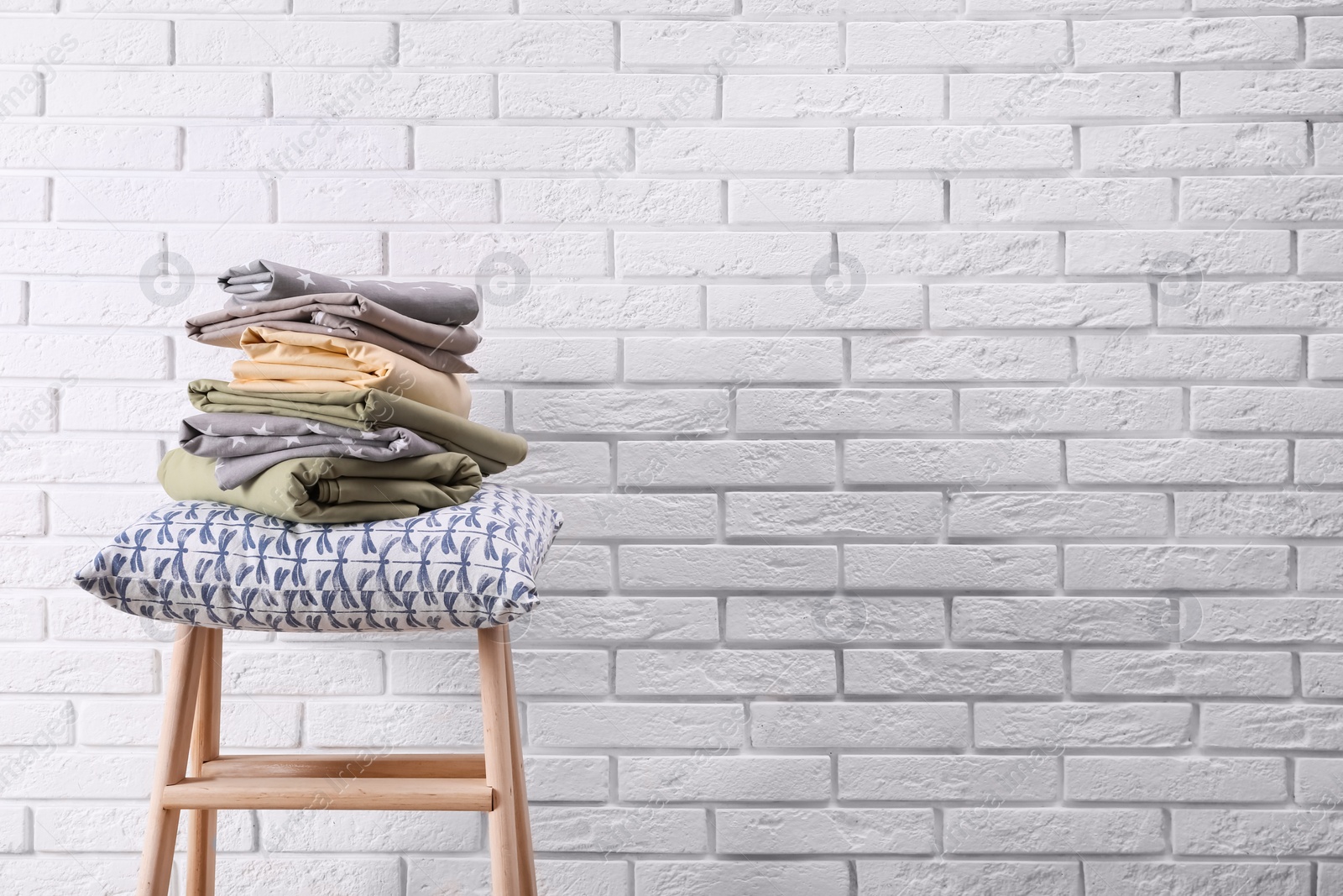 Photo of Stack of clean bed sheets and pillow on stool near white brick wall. Space for text