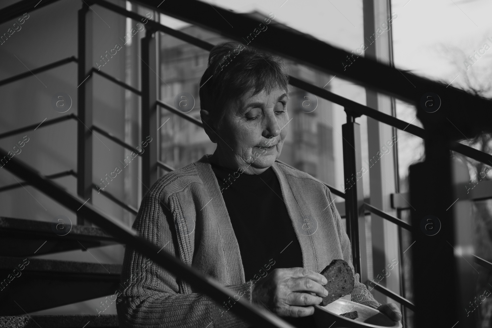 Photo of Poor senior woman with bread sitting on stairs indoors, black and white effect