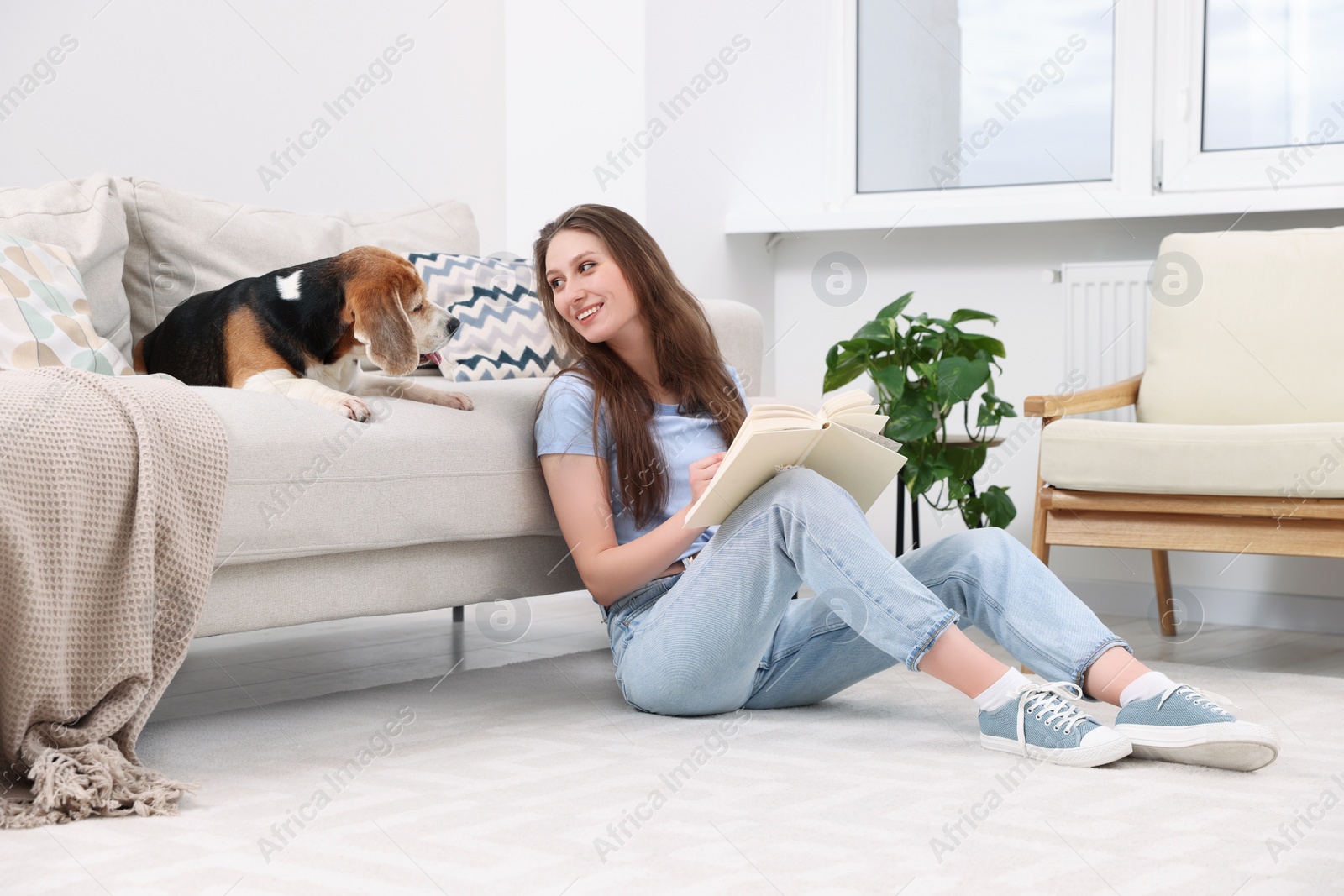 Photo of Happy young woman reading book near her cute Beagle dog on couch at home. Lovely pet