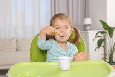 Cute little child eating tasty yogurt from plastic cup with spoon in high chair indoors