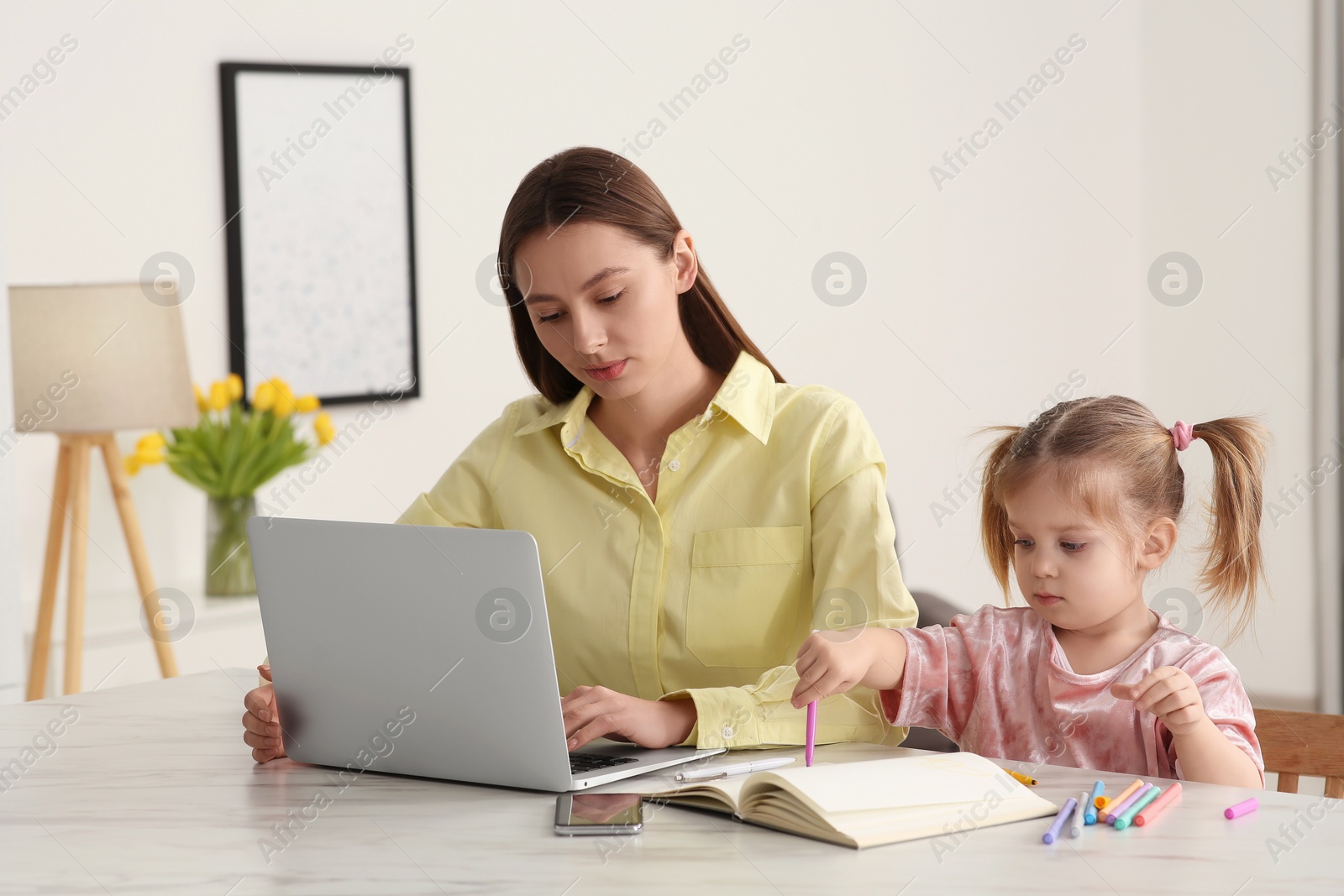 Photo of Woman working remotely at home. Mother using laptop while daughter drawing at desk