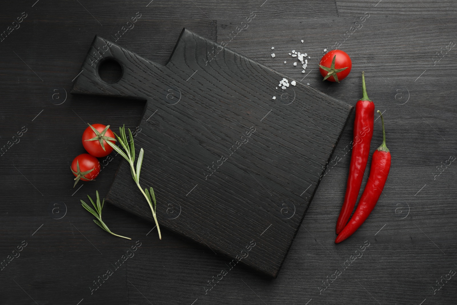 Photo of Cutting board, rosemary, chili peppers and tomatoes on black wooden table, flat lay. Space for text