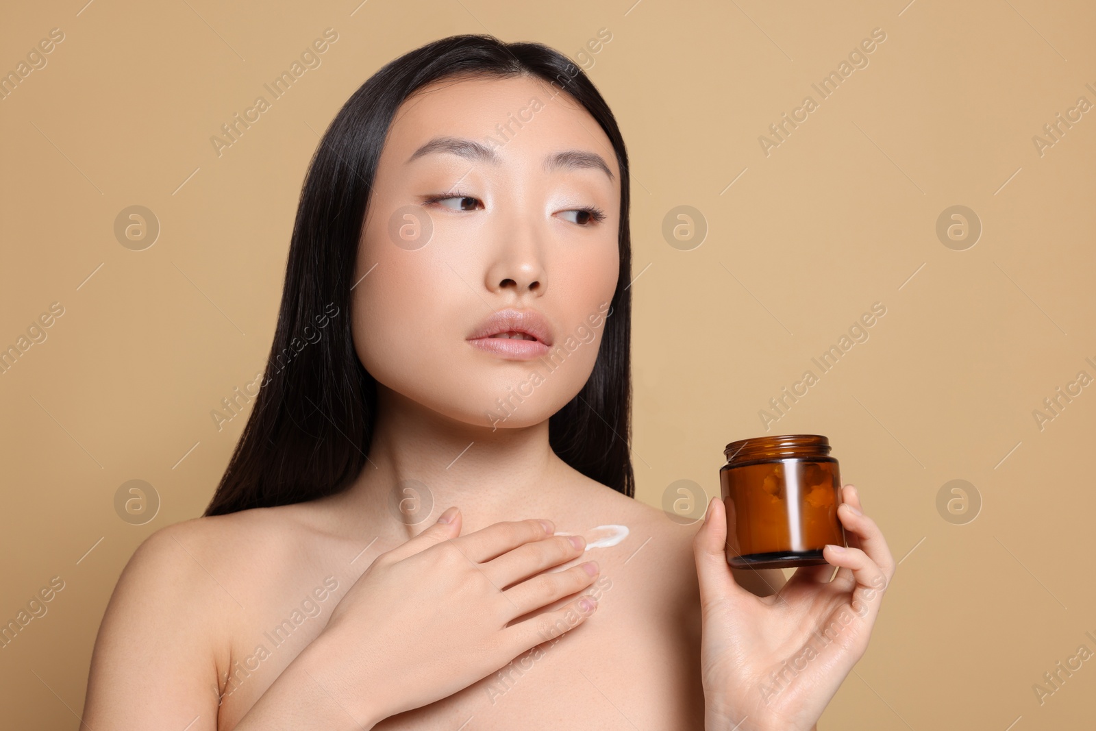 Photo of Beautiful young Asian woman with jar of body cream on beige background