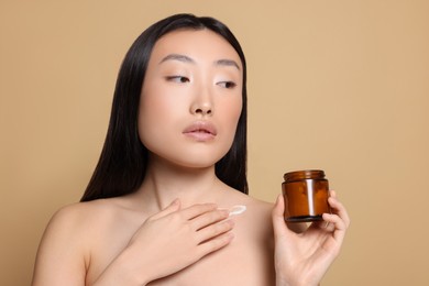 Beautiful young Asian woman with jar of body cream on beige background