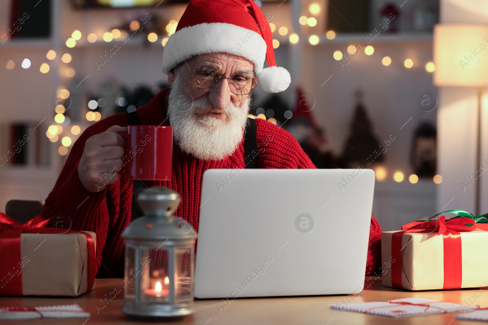 Photo of Santa Claus using laptop and drinking hot beverage at his workplace in room decorated for Christmas