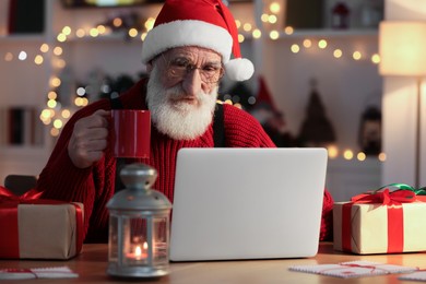 Photo of Santa Claus using laptop and drinking hot beverage at his workplace in room decorated for Christmas
