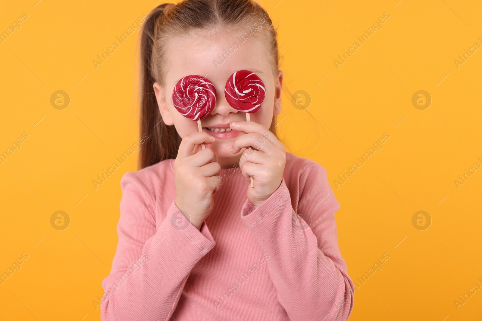 Photo of Happy little girl with bright lollipops covering eyes on orange background