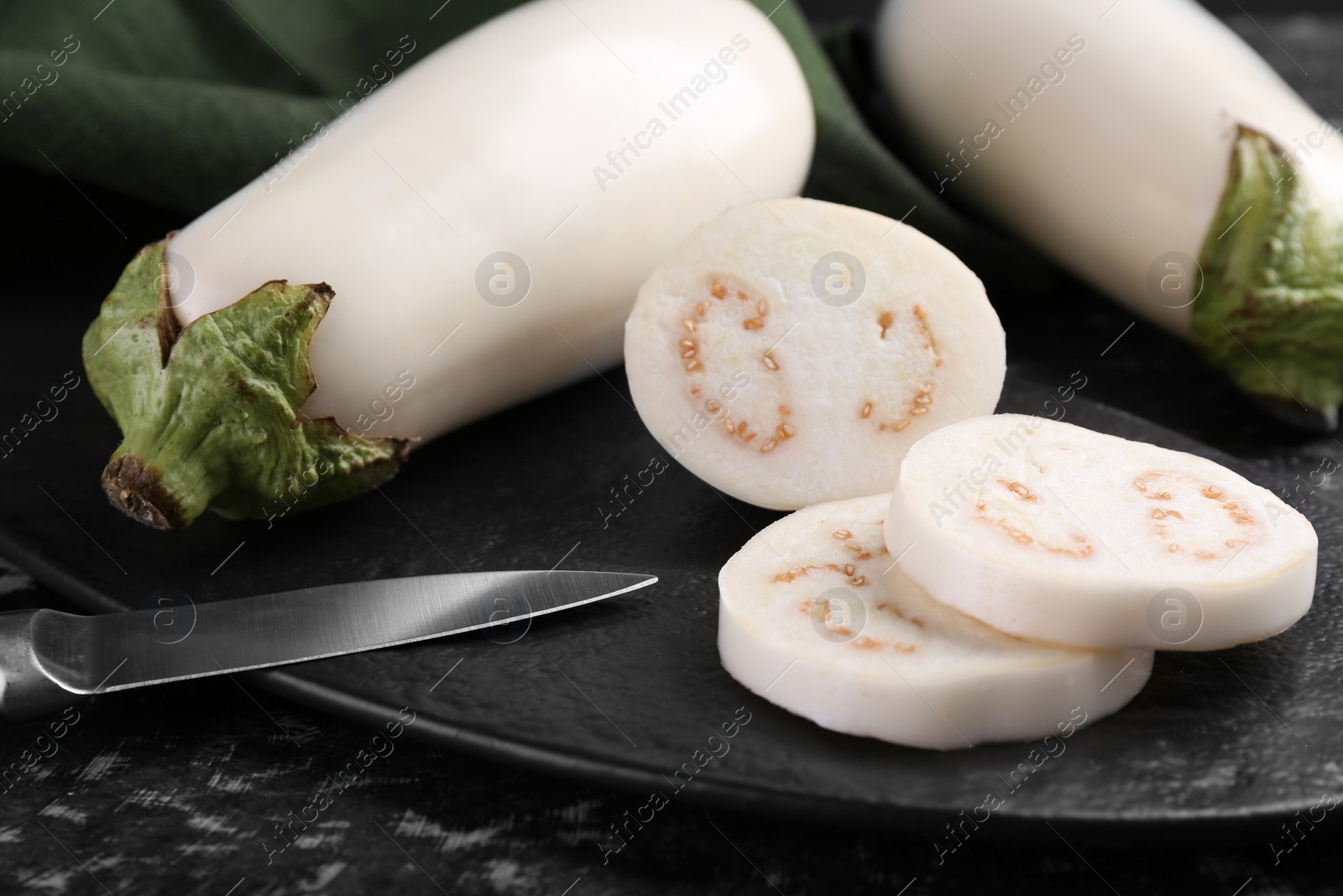 Photo of Board, raw white eggplants and knife on black table, closeup