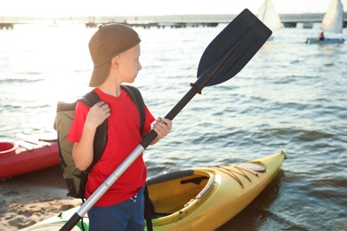 Photo of Little boy with paddle near kayak on river shore. Summer camp activity