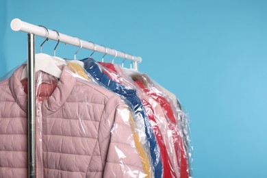 Photo of Dry-cleaning service. Many different clothes in plastic bags hanging on rack against light blue background, space for text