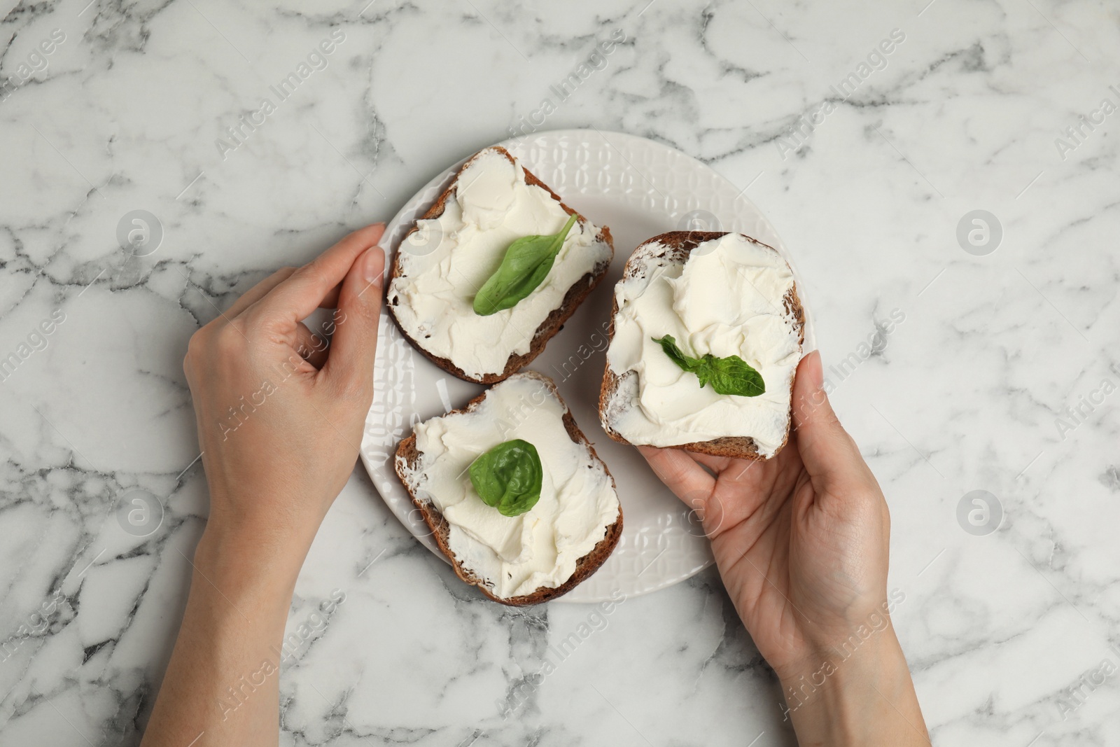 Photo of Woman holding bread with cream cheese at white marble table, top view