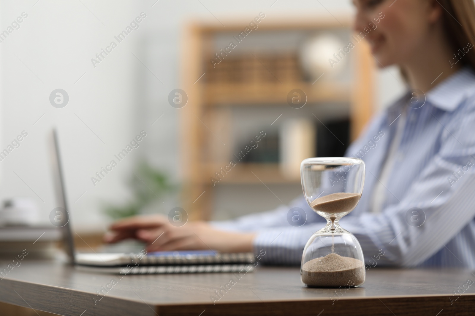 Photo of Hourglass with flowing sand on desk. Woman using laptop indoors, selective focus