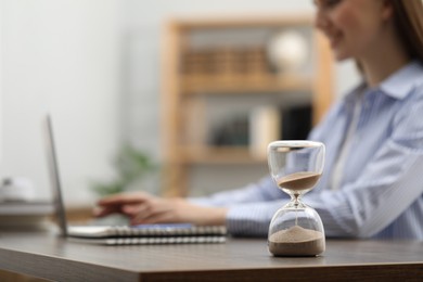 Hourglass with flowing sand on desk. Woman using laptop indoors, selective focus