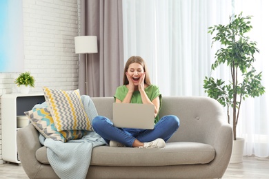 Photo of Emotional young woman with laptop celebrating victory on sofa at home