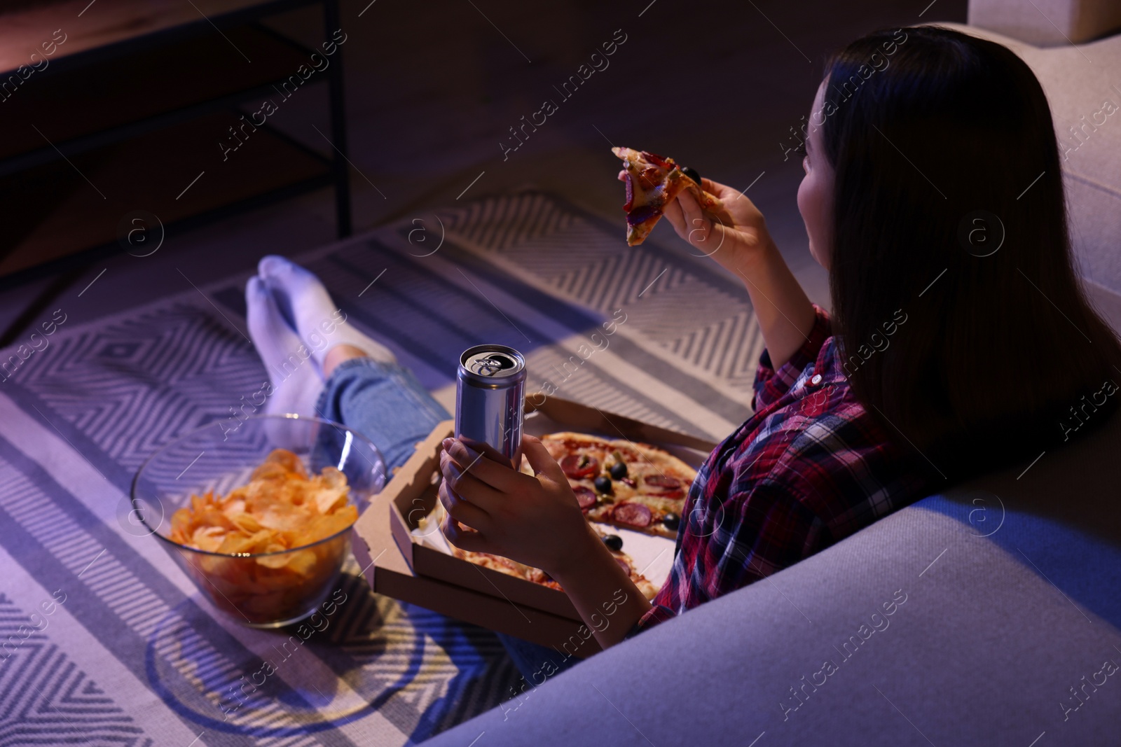 Photo of Young woman eating pizza while watching TV in room at night. Bad habit