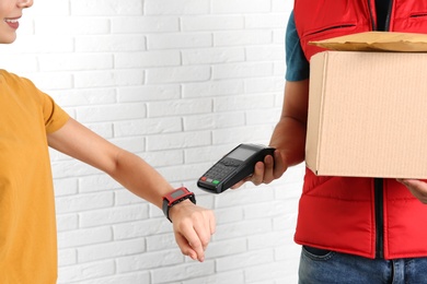 Photo of Young woman with smartwatch using terminal for delivery payment against brick wall, closeup