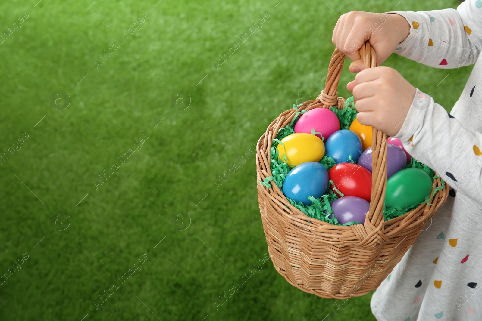 Photo of Little girl with basket full of Easter eggs on green grass, closeup. Space for text