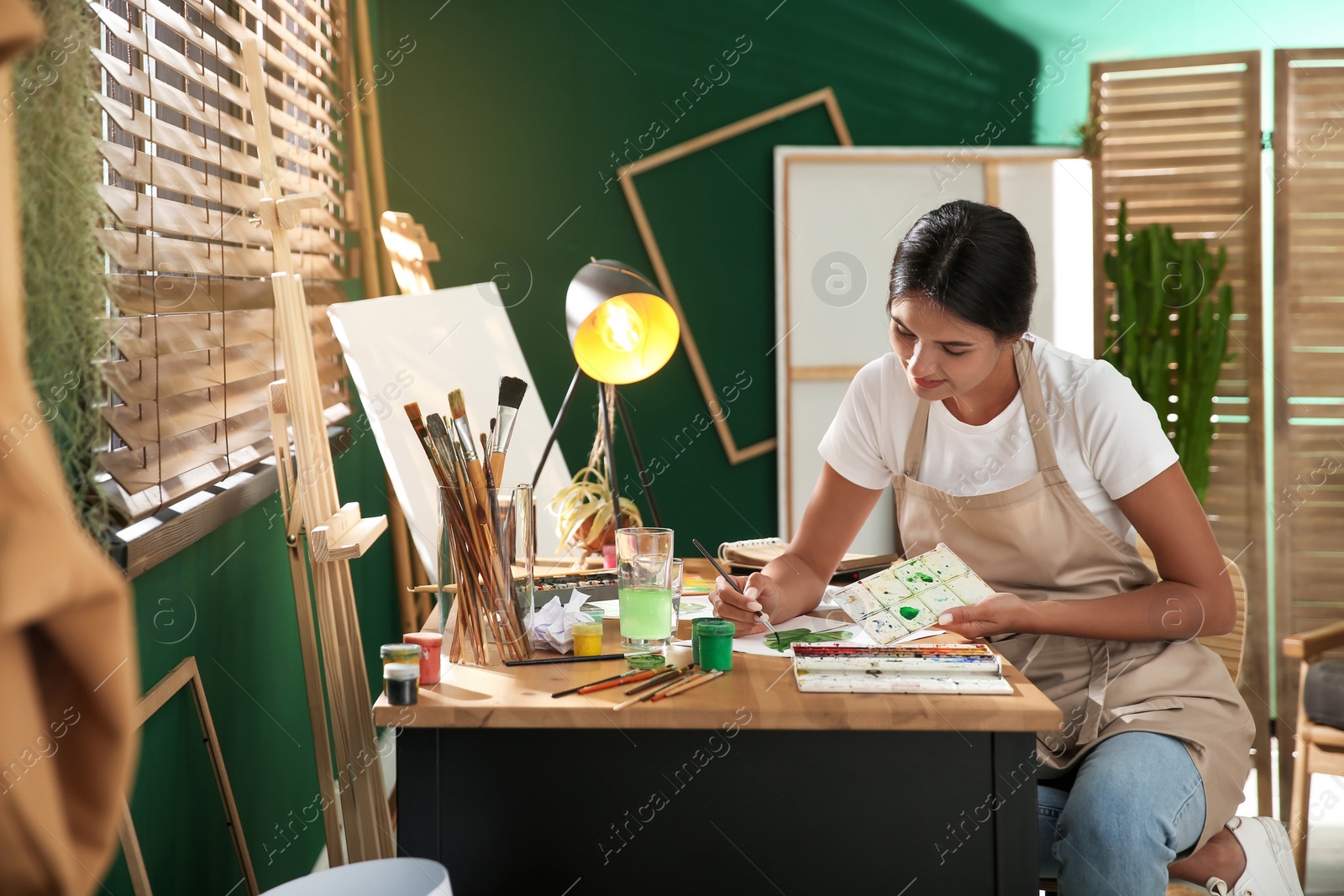 Photo of Young woman drawing leaf with watercolors at table indoors